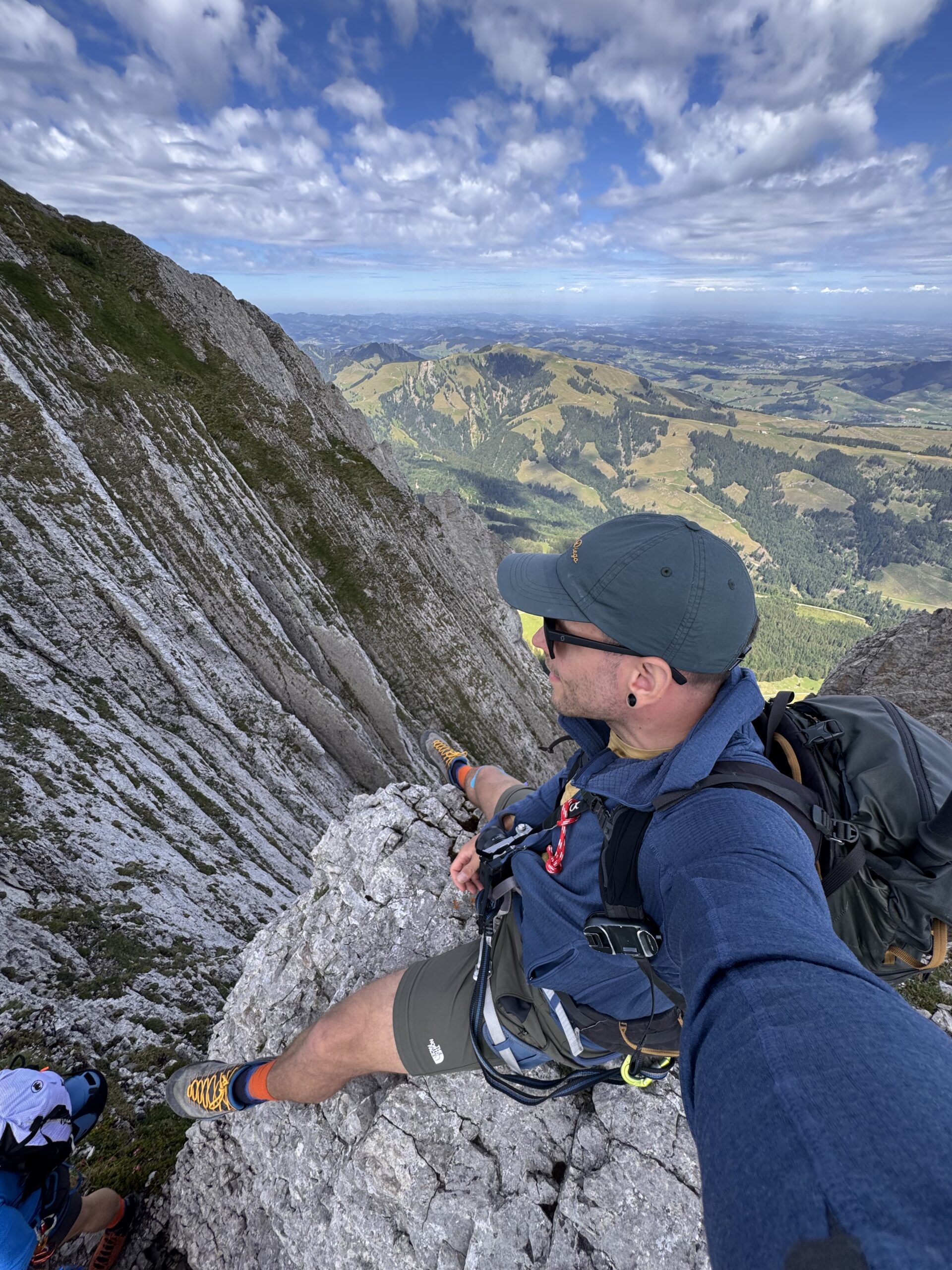 Alex sitzt mit Rucksack am Rand eines schmalen Felsgrats mit Bergpanorama